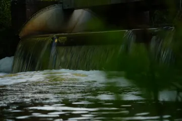 Water spills over a weir in smooth, blurred streaks, seen through soft green leaves.