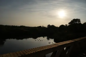 The sun behind thin cloud above the dark lake, with the edge of the bridge in the corner.
