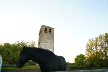 A dark horse stands beside the fence in front of the old brick tower and sunlit trees.