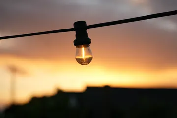 A glowing bulb hangs from a black cable against a softly blurred orange sunset and dark rooftops.